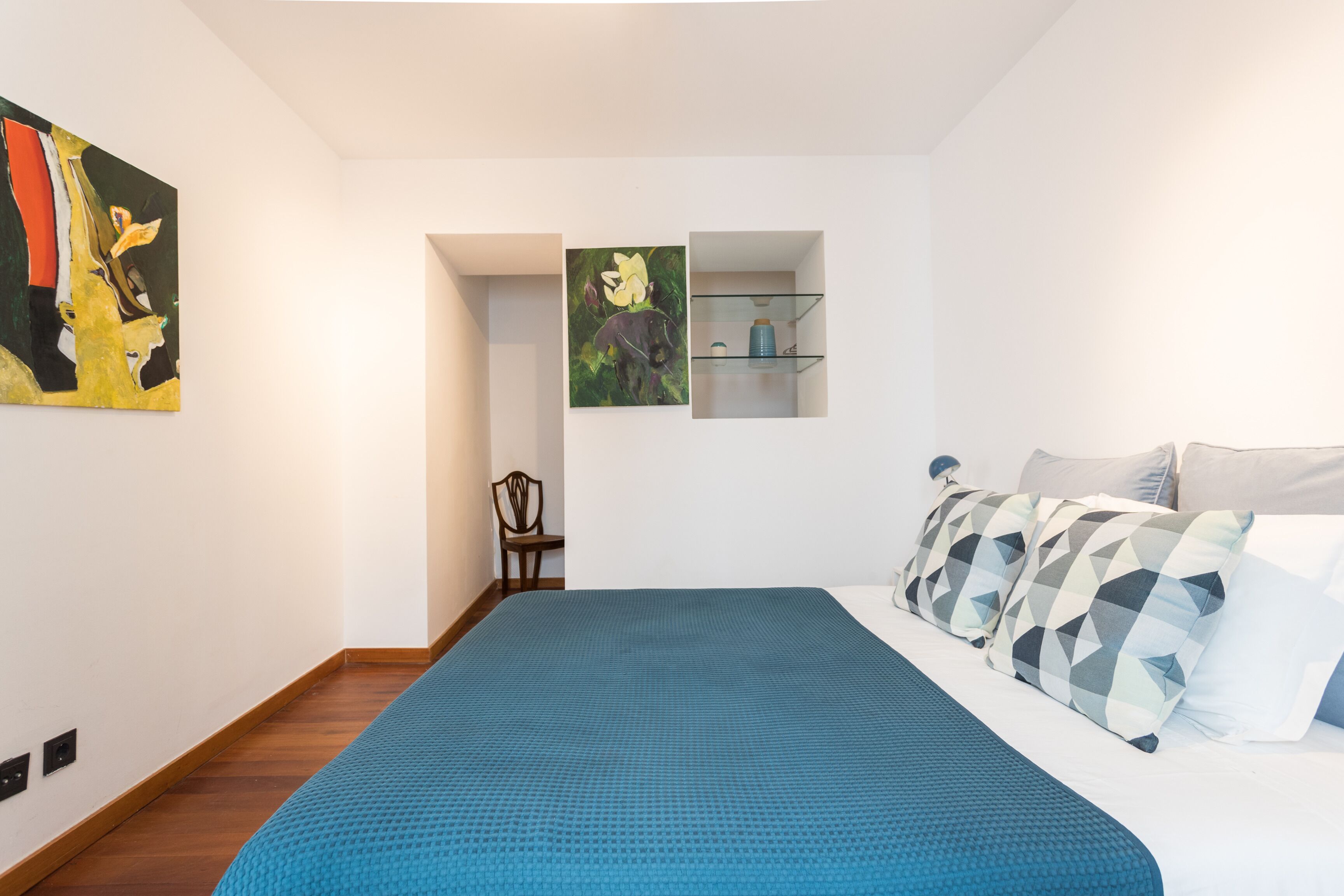 Un dormitorio moderno con una cama grande, una colcha azul y dos estantes empotrados con adornos decorativos.