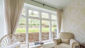Una habitación con un ventanal grande, una silla de mimbre, un sillón mullido y una pequeña mesa de madera.