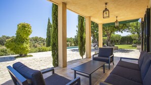 Eine Terrasse im Freien mit überdachter Sitzgelegenheit, inklusive Tisch und Stühlen, mit Blick auf einen angelegten Garten und Poolbereich.
