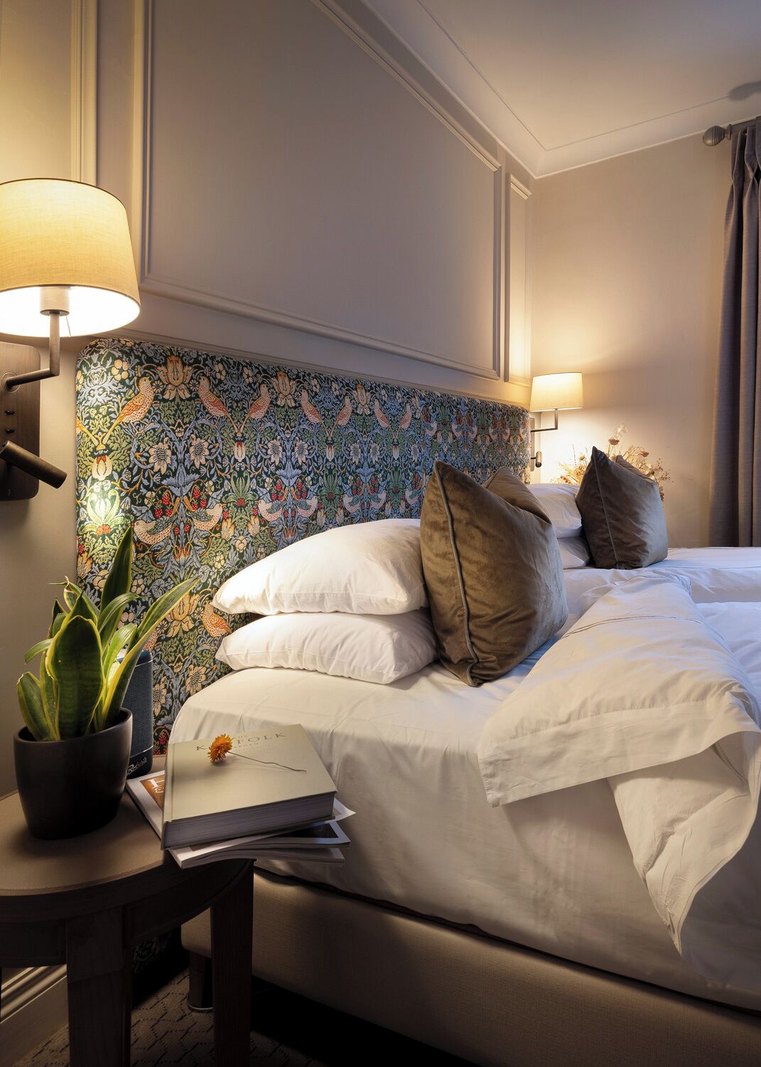 A hotel room with a bed, two bedside lamps, a potted plant, and a decorative headboard.
