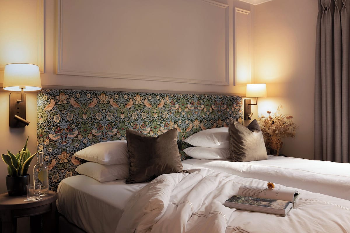 A hotel room with a bed, two wall-mounted lamps, a potted plant, and a book on the bed.