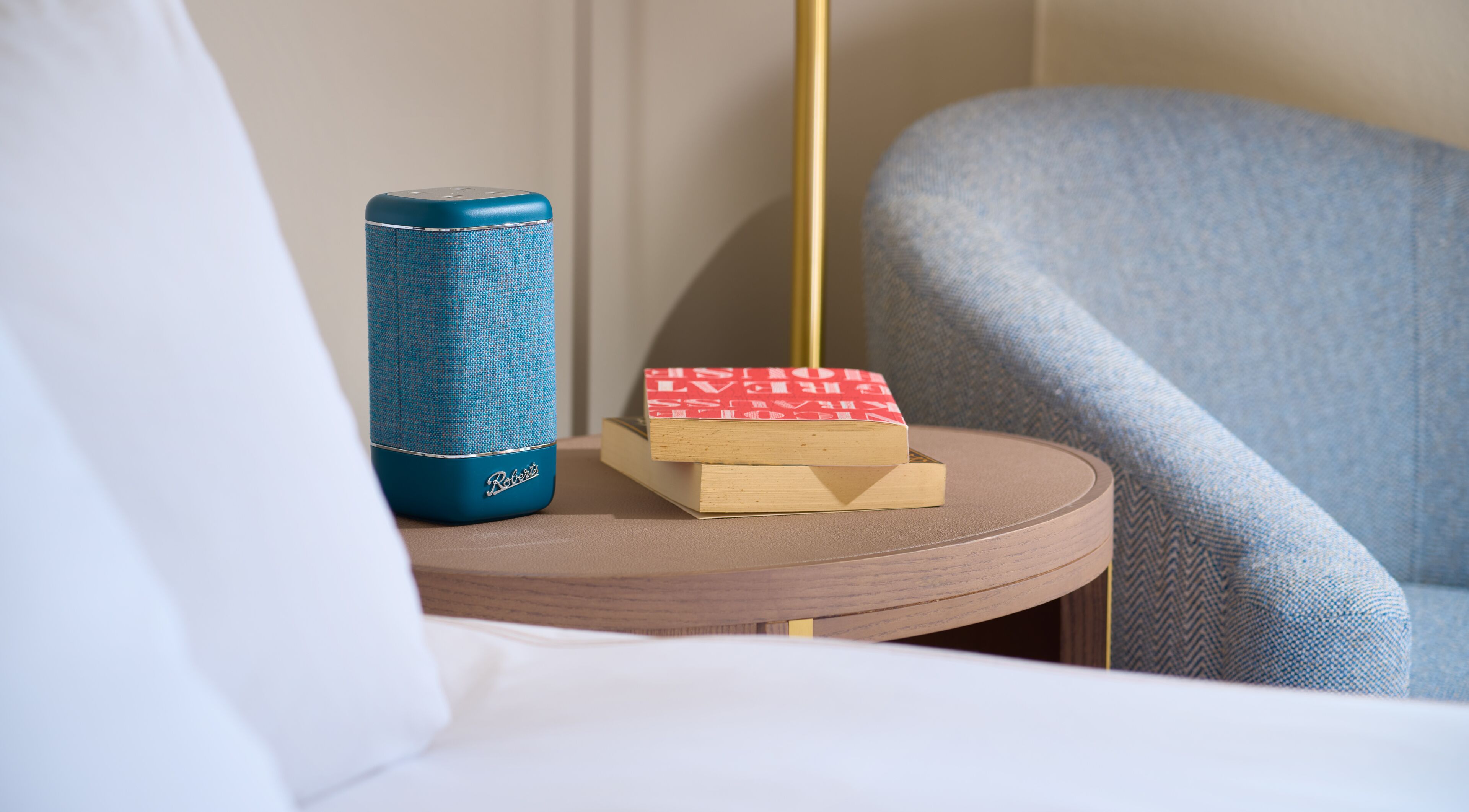 Una habitación de hotel con una mesita redonda de madera, un altavoz azul, un libro rojo y un sillón gris.
