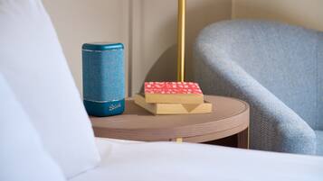 A hotel room with a round wooden side table, a blue speaker, a red book, and a grey armchair.