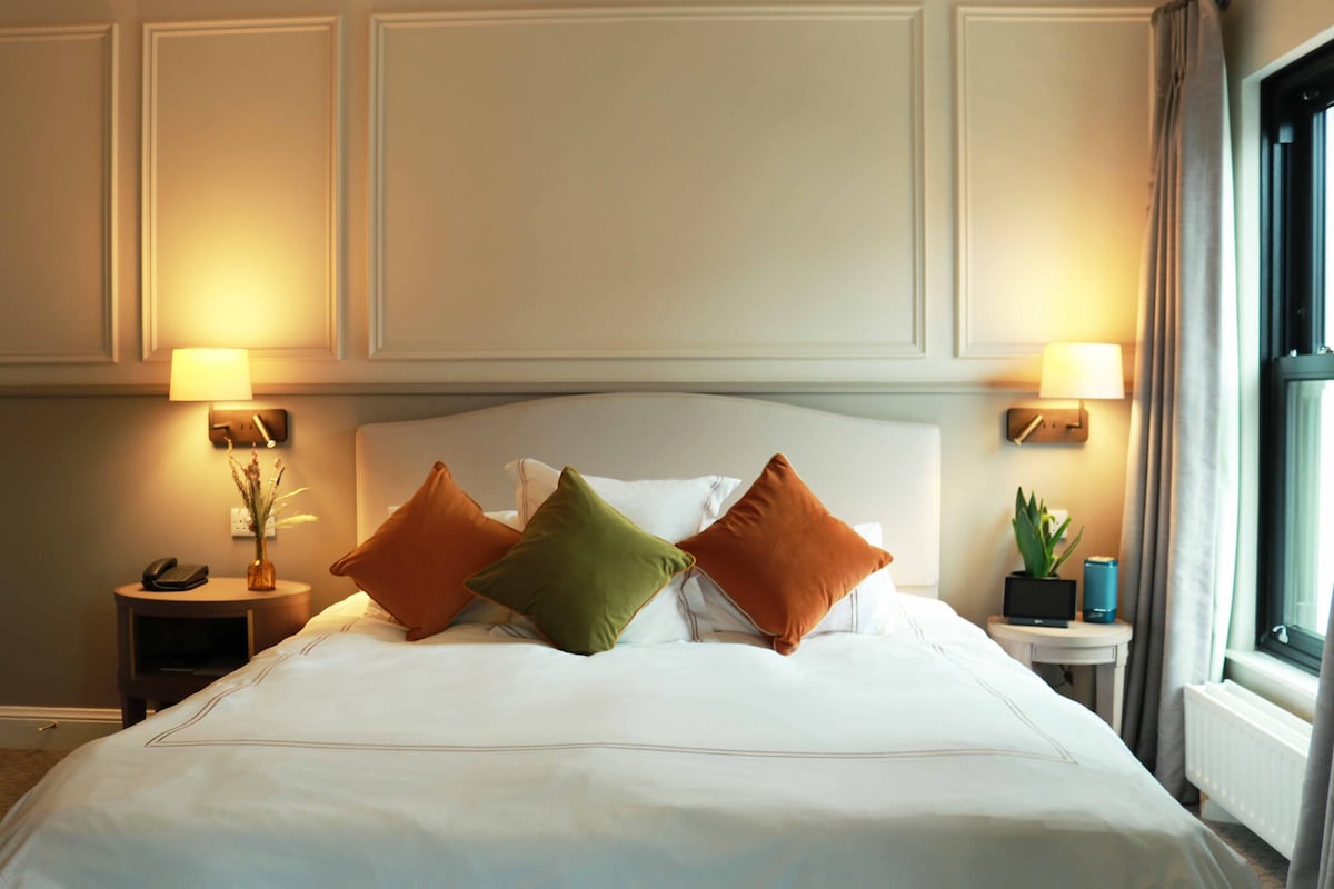 A neatly made bed with white linens and three pillows in different colors. There are two bedside tables, one with a lamp and the other with a plant. The room has a window with curtains.