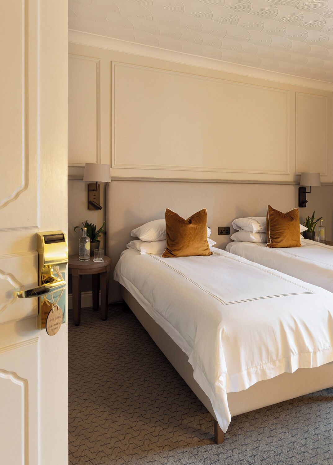 A hotel room with a double bed, a bedside table with a lamp, a small round table with a plant, and a wall-mounted safe.