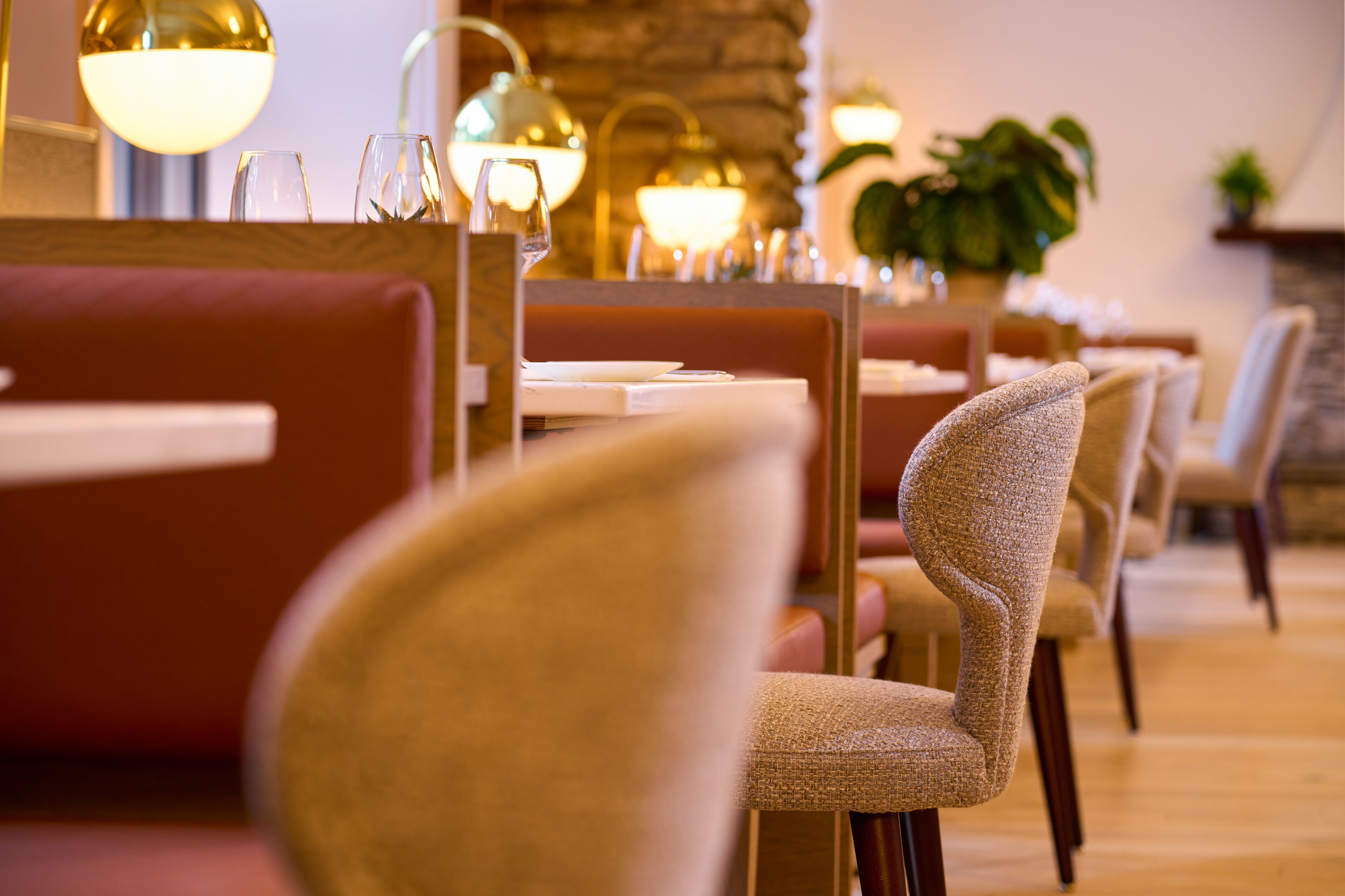 L’intérieur d’un restaurant présente des tables en bois et des chaises rembourrées, un éclairage chaleureux et un mur en pierre en arrière-plan.