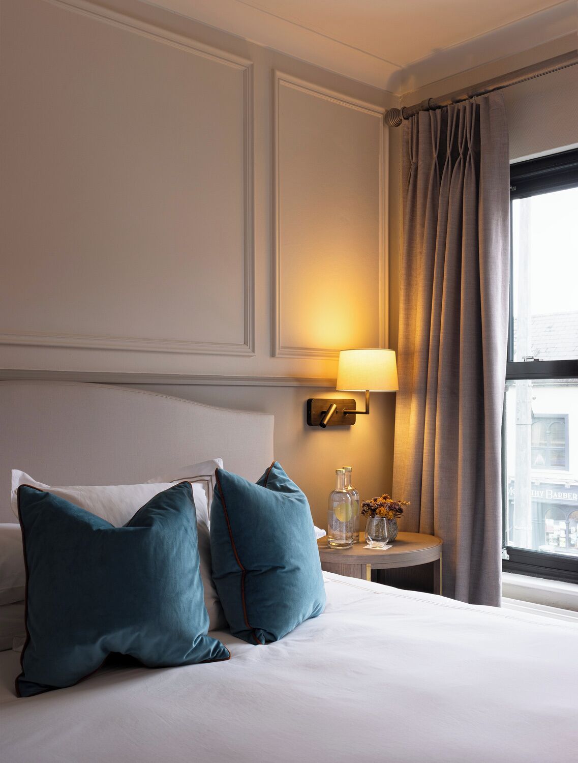 Una habitación de hotel con cama, almohadas, mesita de noche con lámpara y ventana con cortinas.