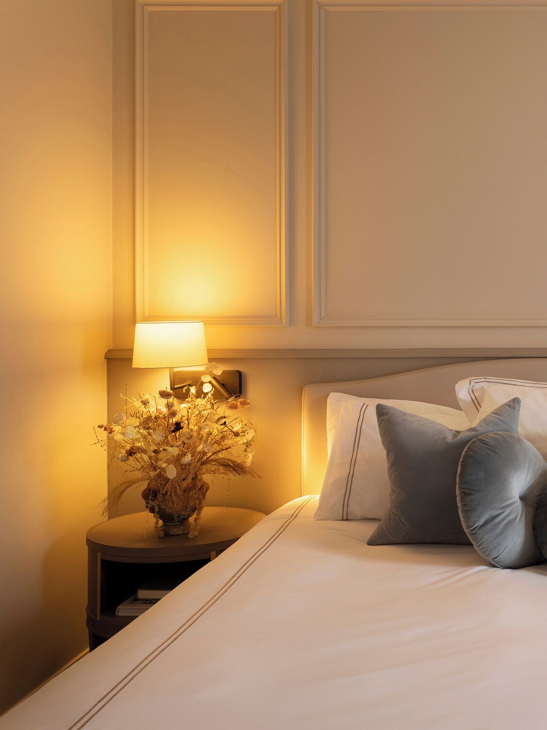 A neatly made bed with pillows, a bedside lamp, and a floral arrangement on a bedside table.