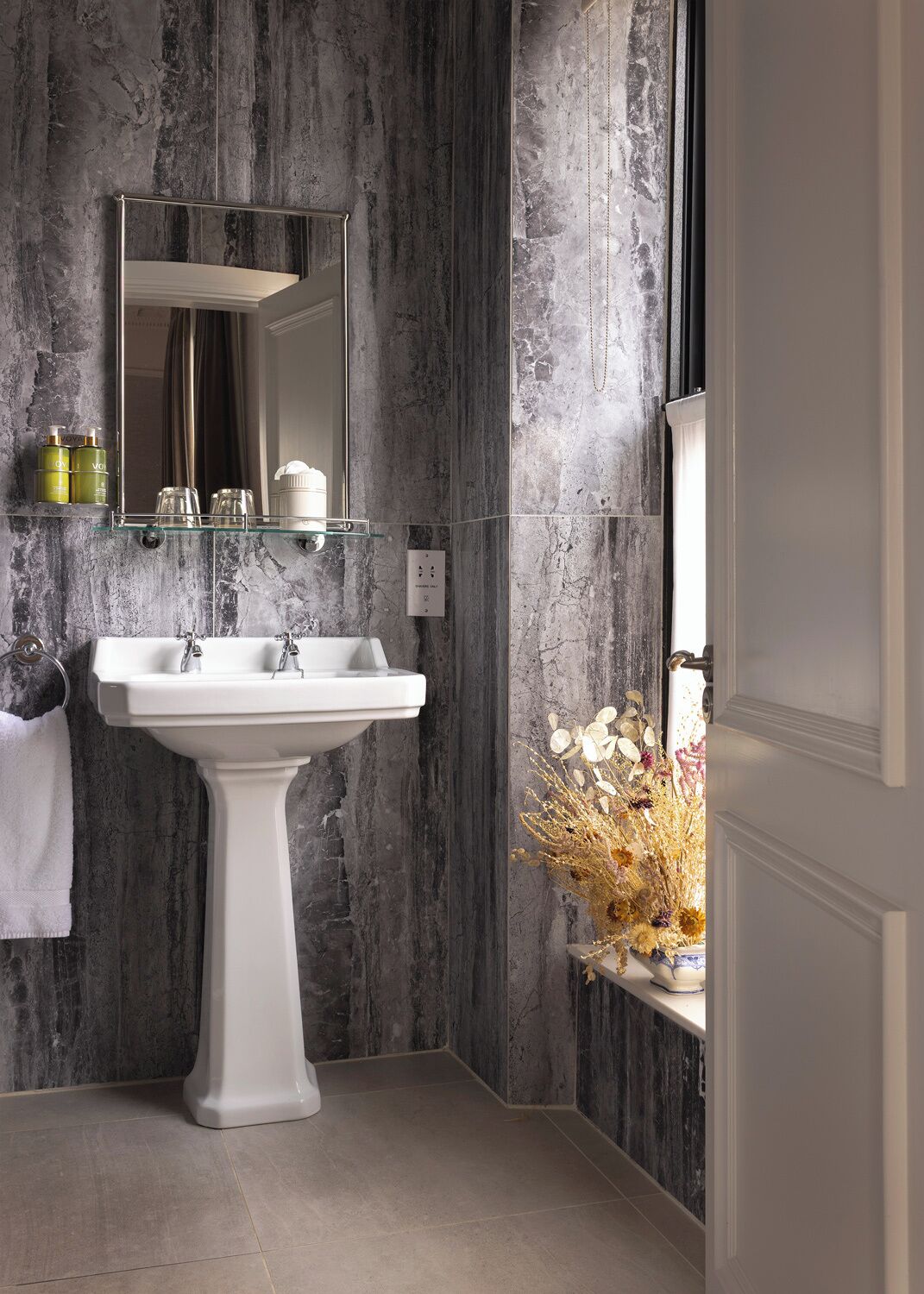 Un baño con un lavamanos blanco de pedestal, un espejo encima y un florero con flores secas en un estante.
