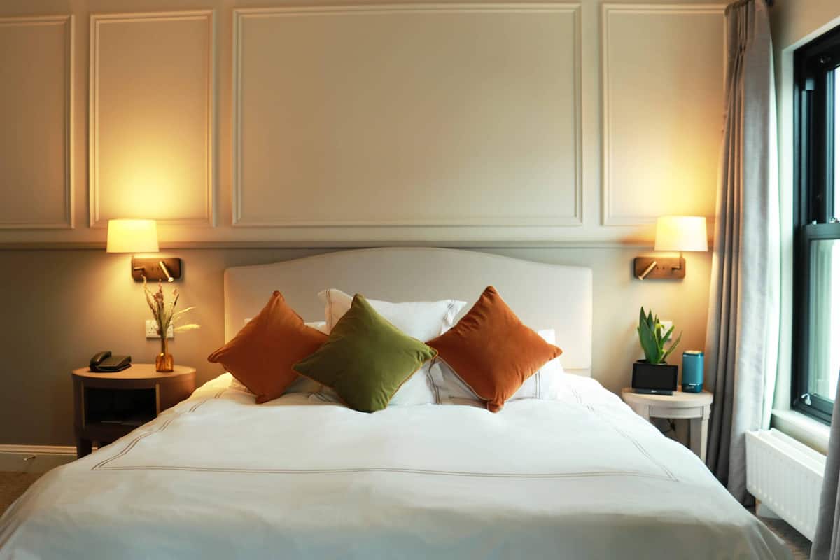 A neatly made bed with white linens and three pillows in different colors. There are two bedside tables, one with a lamp and the other with a plant. The room has a window with curtains.