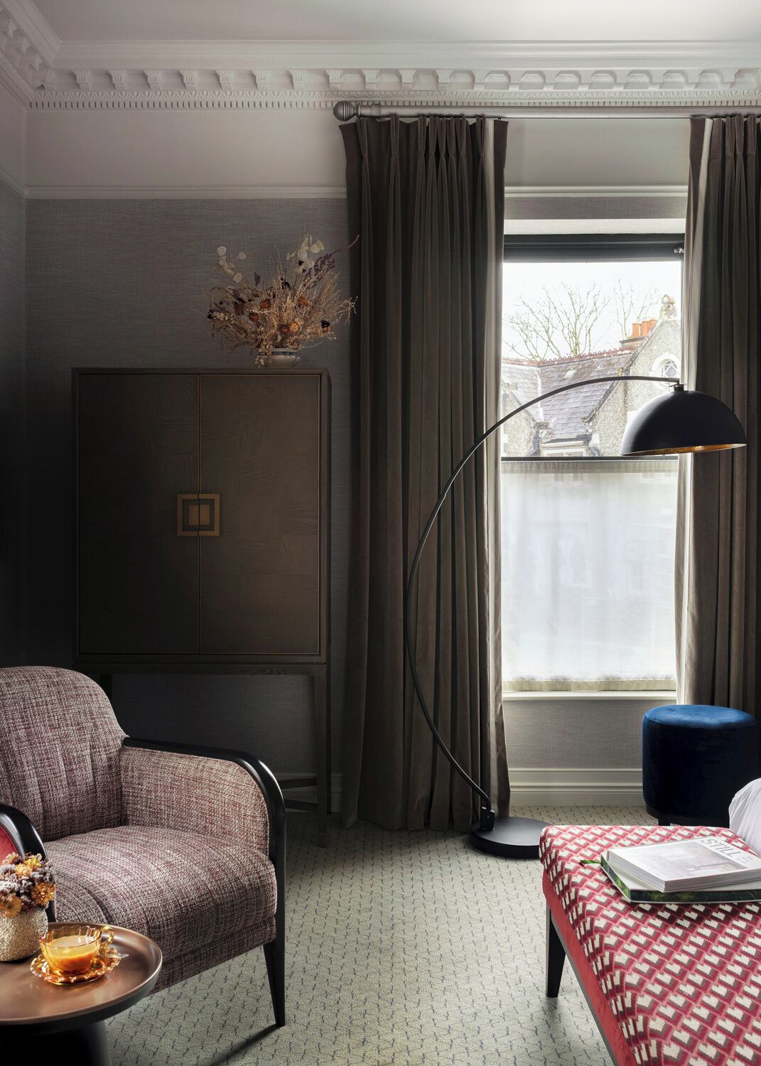 A room with a dark wood wardrobe, a red and white checkered bench, a floor lamp, and a view of a building outside.