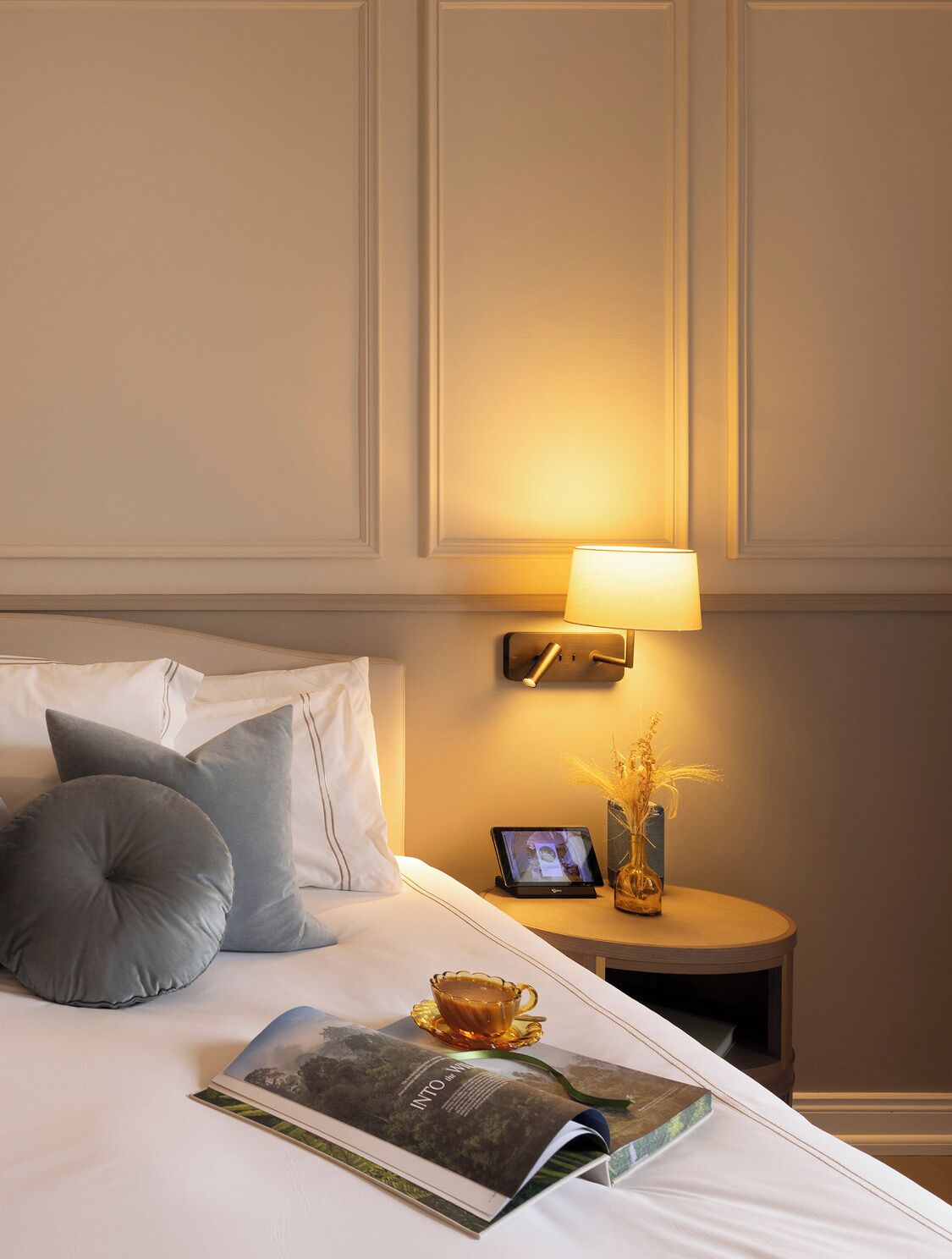 A neatly made bed with pillows, a bedside table with a lamp, a tablet, a vase with dried plants, and a magazine.