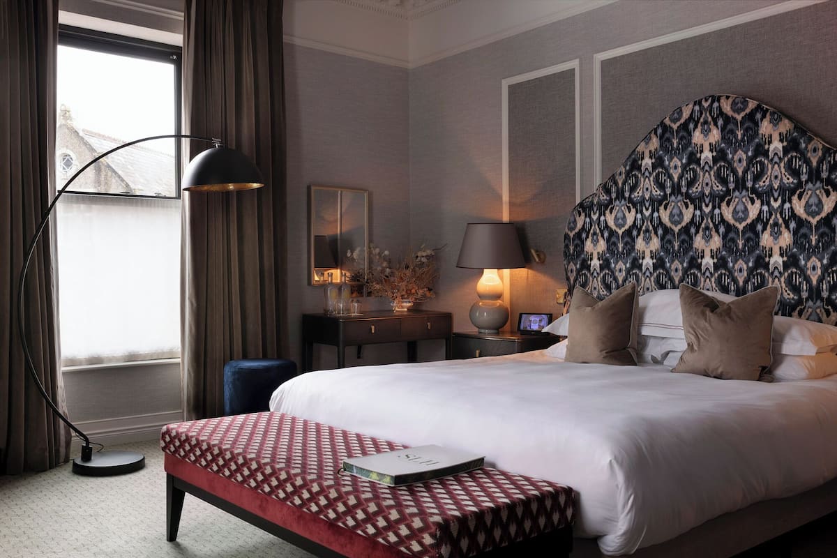 A hotel room with a large bed, a bedside table with a lamp, a small desk, a chair, and a window with curtains.