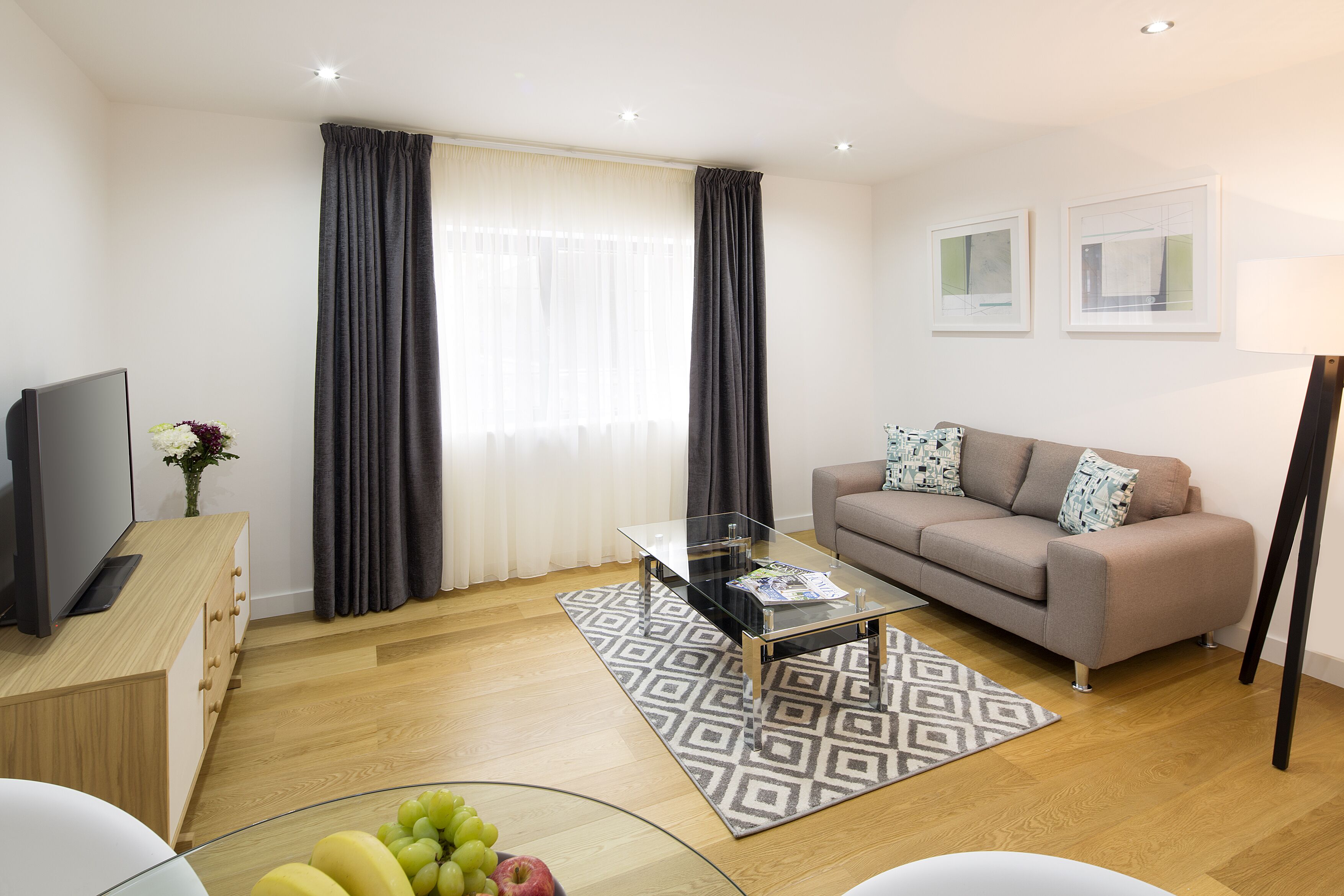 A modern living room with a sofa, a glass coffee table, a TV on a wooden cabinet, and a fruit bowl.