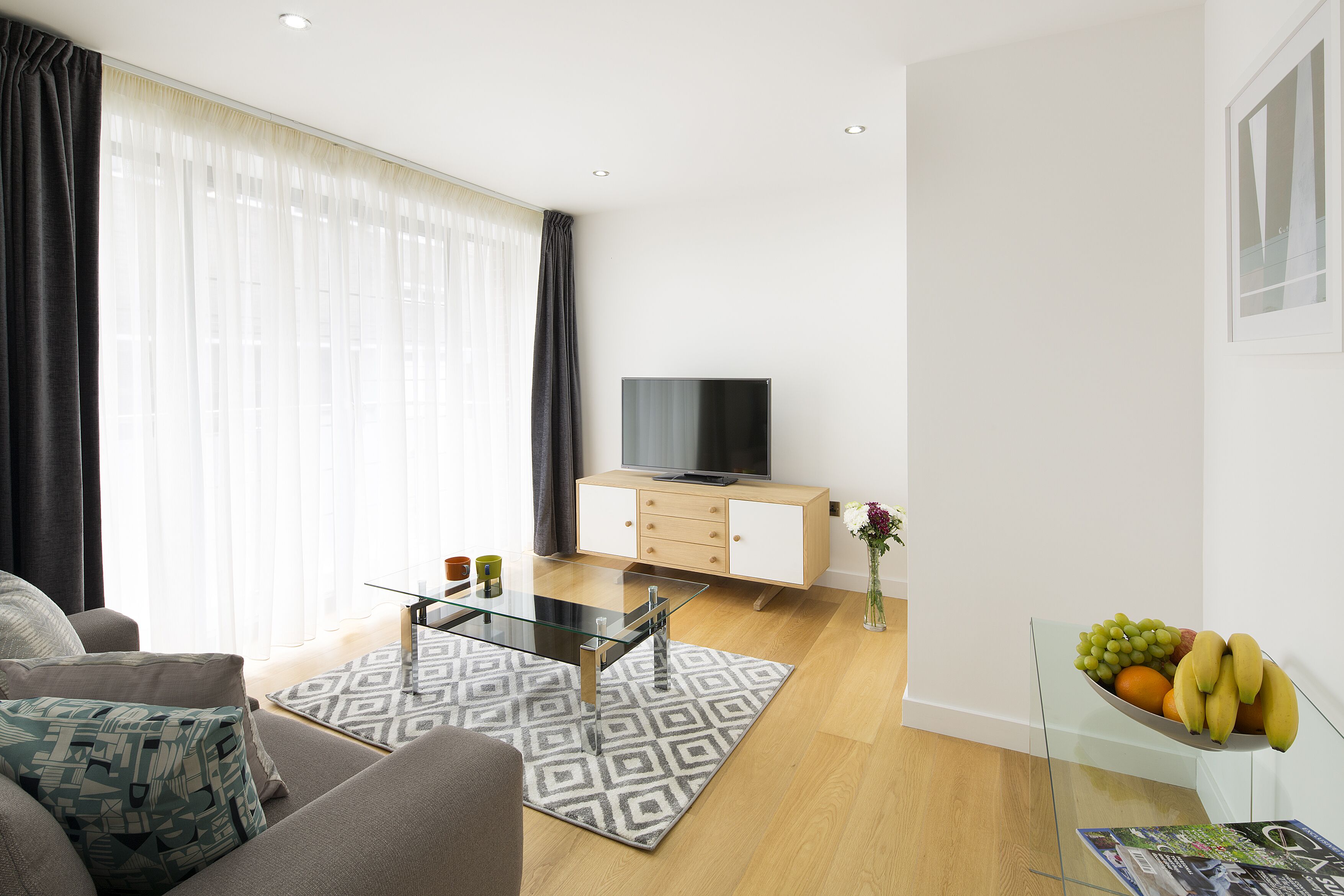 Un salon moderne avec un canapé, une table basse en verre, un téléviseur posé sur un meuble en bois et un panier de fruits sur un présentoir en verre.