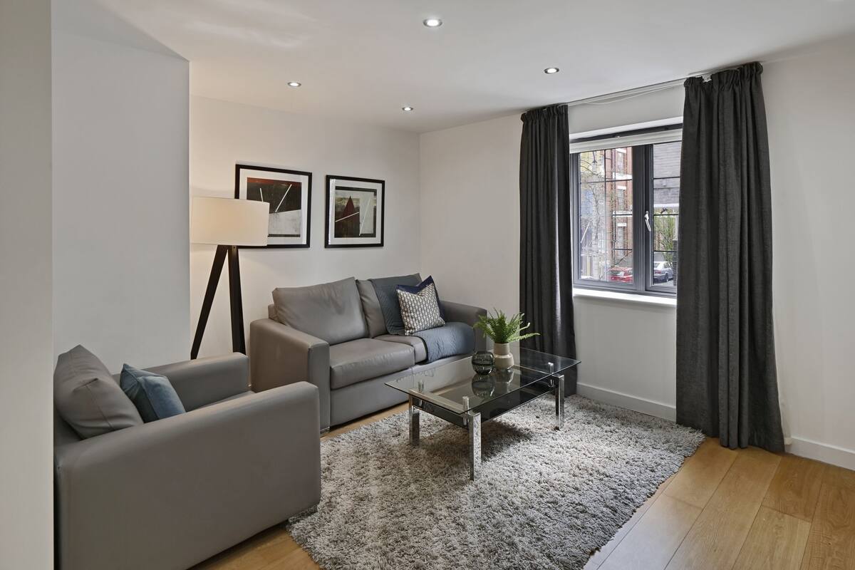 A modern living room with a grey sofa, a glass coffee table, and a large window with curtains.