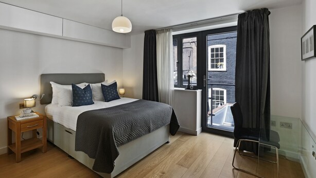 A modern hotel room with a bed, bedside table, lamp, and a view of the outside through a window.