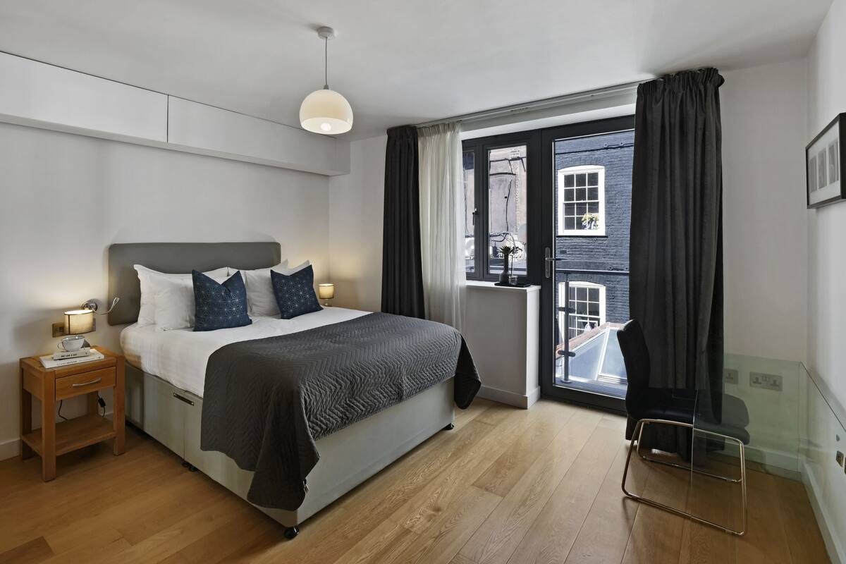 A modern hotel room with a bed, bedside table, lamp, and a view of the outside through a window.