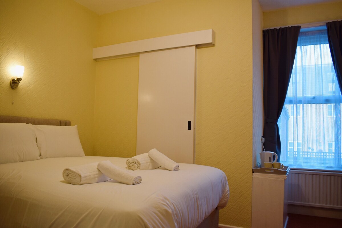 A hotel room with a bed, white linens, a nightstand with a cup and saucer, and a window with curtains.