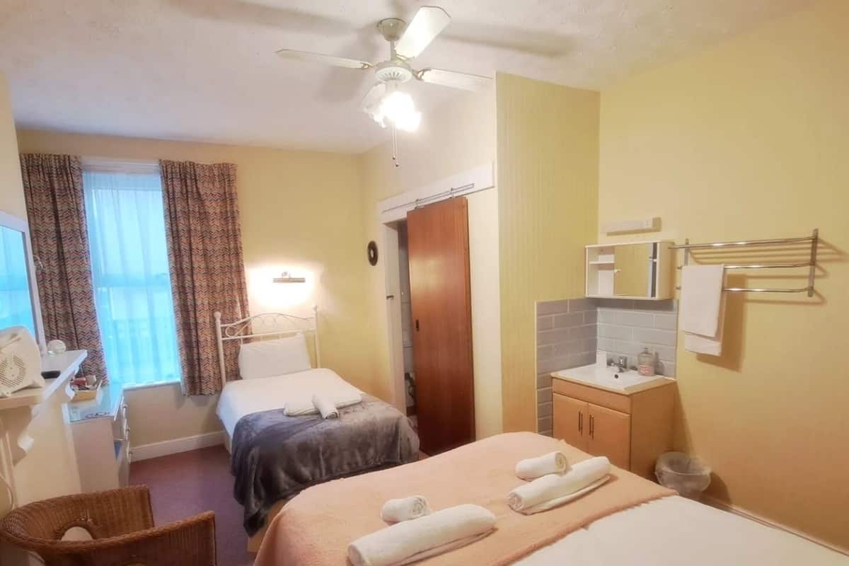 A hotel room with two beds, a desk with a chair, a ceiling fan, a window with curtains, and a bathroom with a sink and towel rack.
