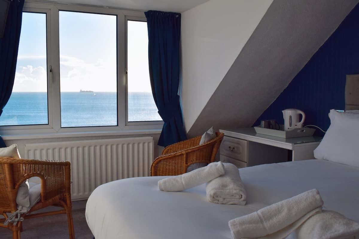 A hotel room with a large bed, a view of the sea, a window with curtains, a wicker chair, a bedside table with a kettle, and a white radiator.
