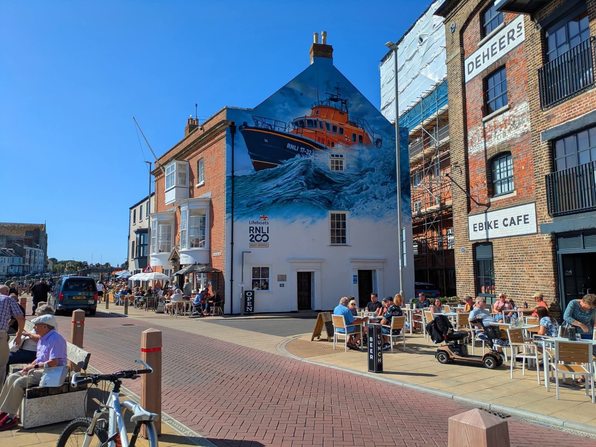 A building with a mural of a lifeboat on the side, a sign for 'Lifeboats East Street,' and a 'Bike Cafe' adjacent to it.