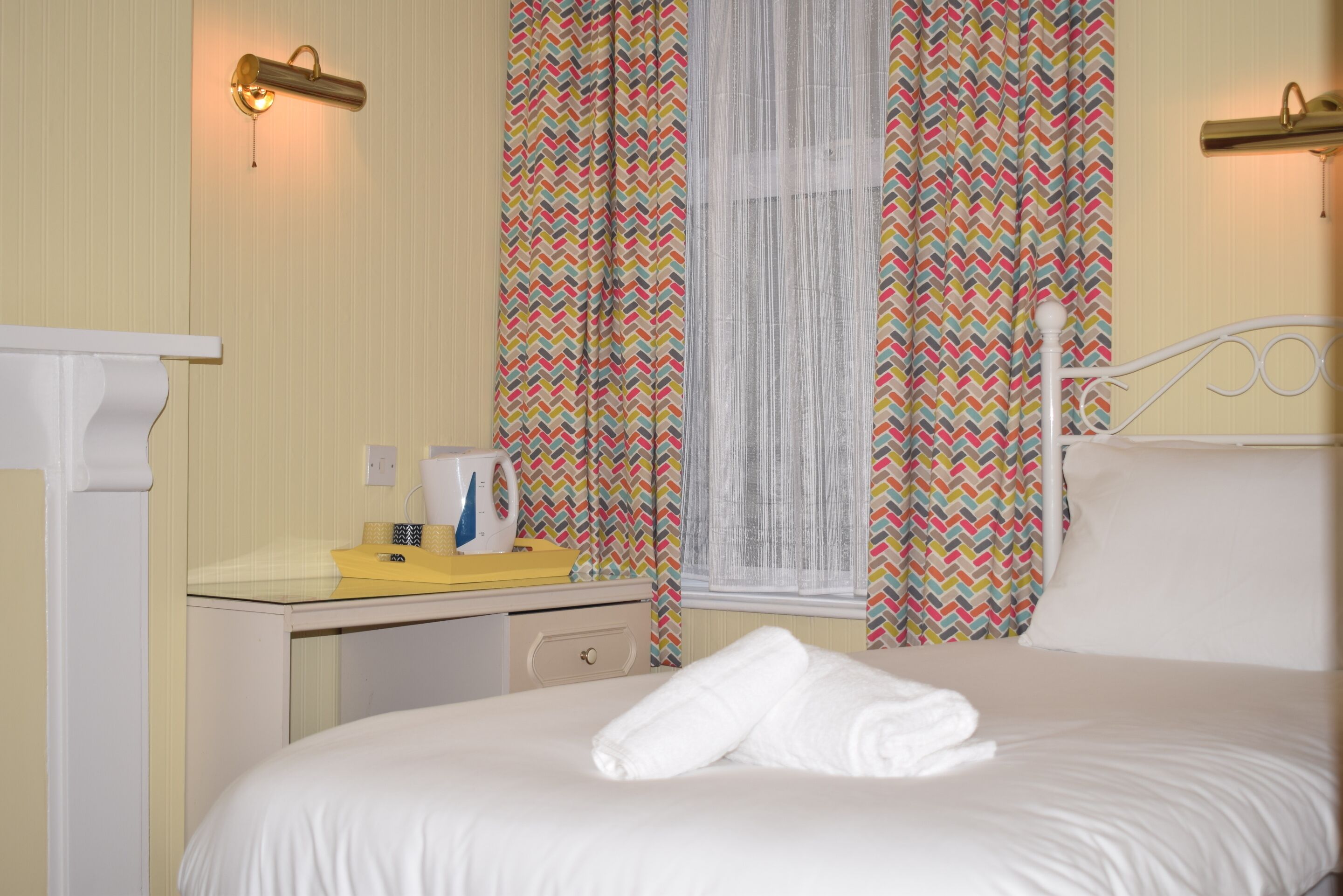 Una cama bien hecha con sábanas blancas, una mesita de noche con un objeto amarillo y una ventana con cortinas estampadas.
