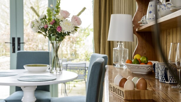 A dining area with a round table, chairs, a vase with flowers, a bowl, a fruit bowl, a lamp, and a wooden box labeled "DOZEN". - The Hall - luxury barn conversion in the Cotwolds (Bristol)