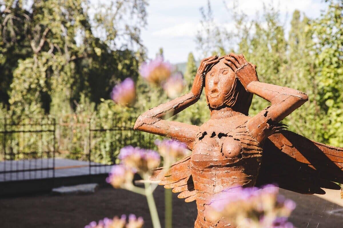 Una scultura raffigura una figura con le braccia aperte, sullo sfondo di alberi e fiori viola.