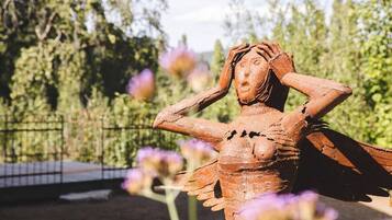 Una scultura raffigura una figura con le braccia aperte, sullo sfondo di alberi e fiori viola.