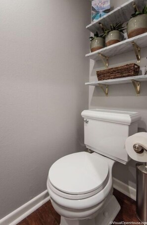 Een badkamer met een wit toilet, een witte wastafel en een witte douche. Boven het toilet staan planken met potplanten en een mand.