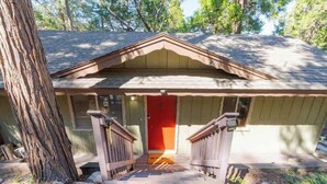 Une maison en bois avec une porte rouge, une véranda dotée de balustrades en bois et un arbre sur la gauche.