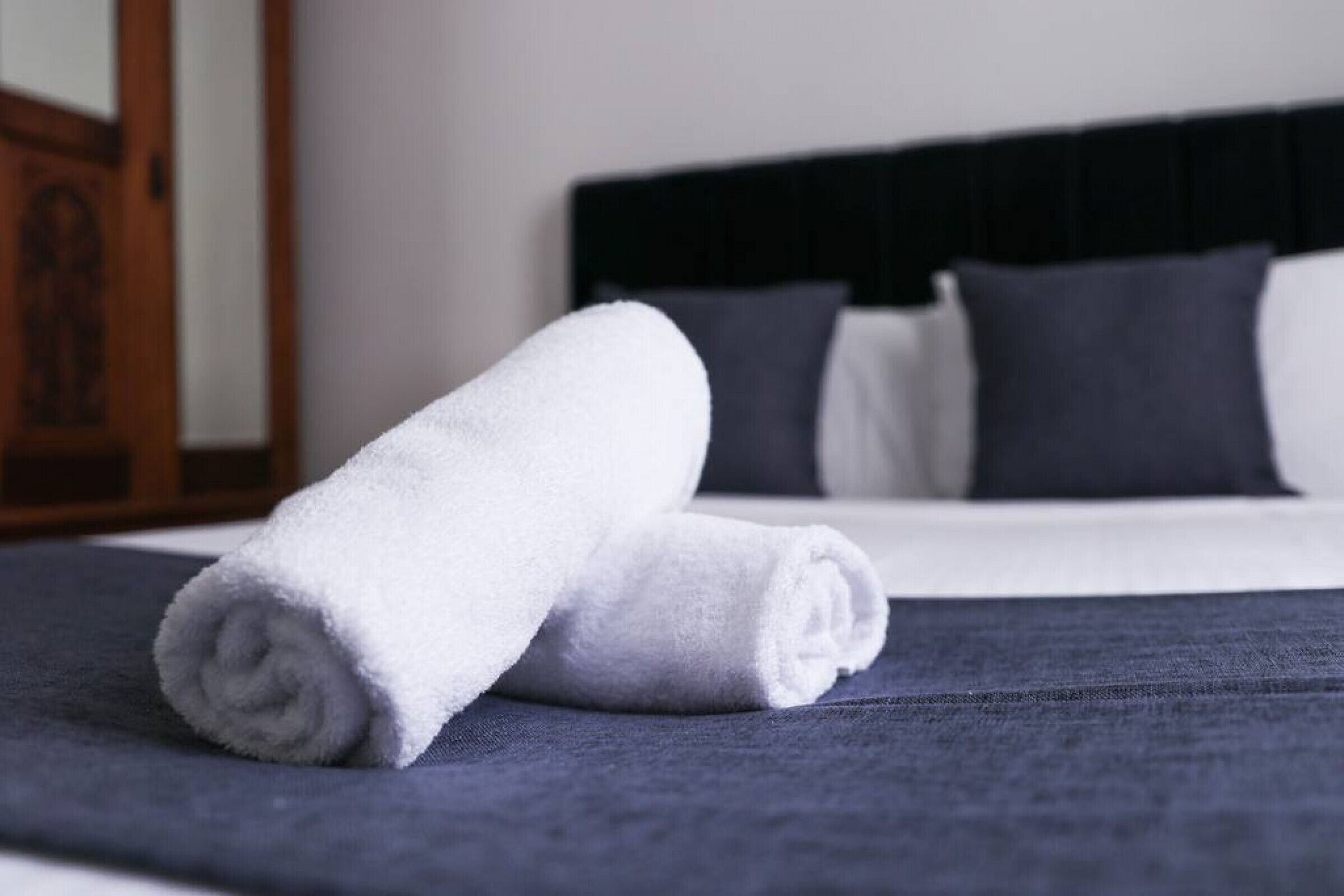 A neatly made bed with a dark blue bedspread and two rolled white towels.