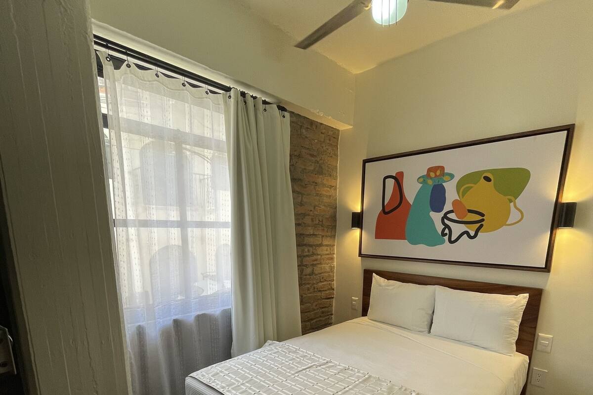 Une chambre d’hôtel avec un lit, un ventilateur de plafond, une fenêtre avec des rideaux, un mur en briques et un tableau accroché au mur.