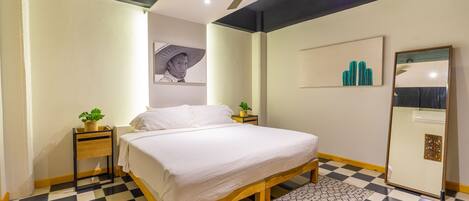 Un dormitorio con piso de cuadros, una cama con ropa blanca, un ventilador de techo, una mesita de noche con una planta y un espejo en la pared.