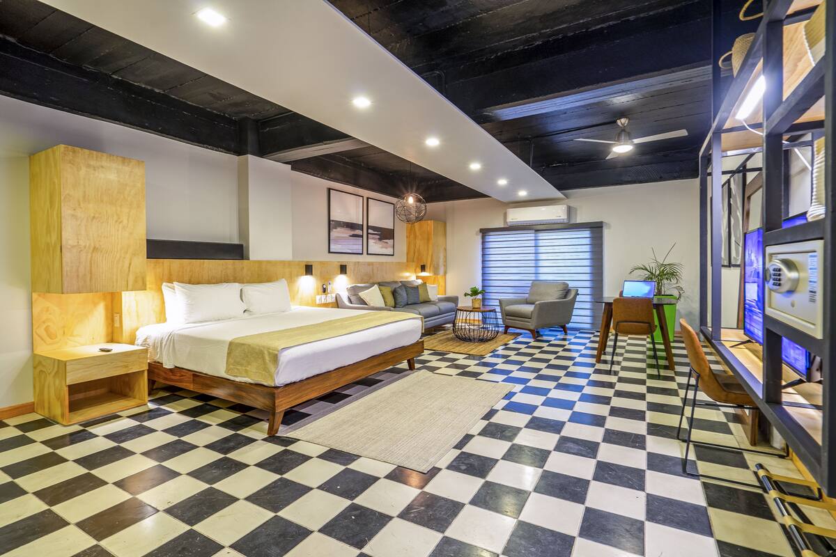 Une chambre d’hôtel avec un sol à carreaux, un lit avec une tête de lit en bois, un bureau avec un ordinateur et une fenêtre avec des stores.