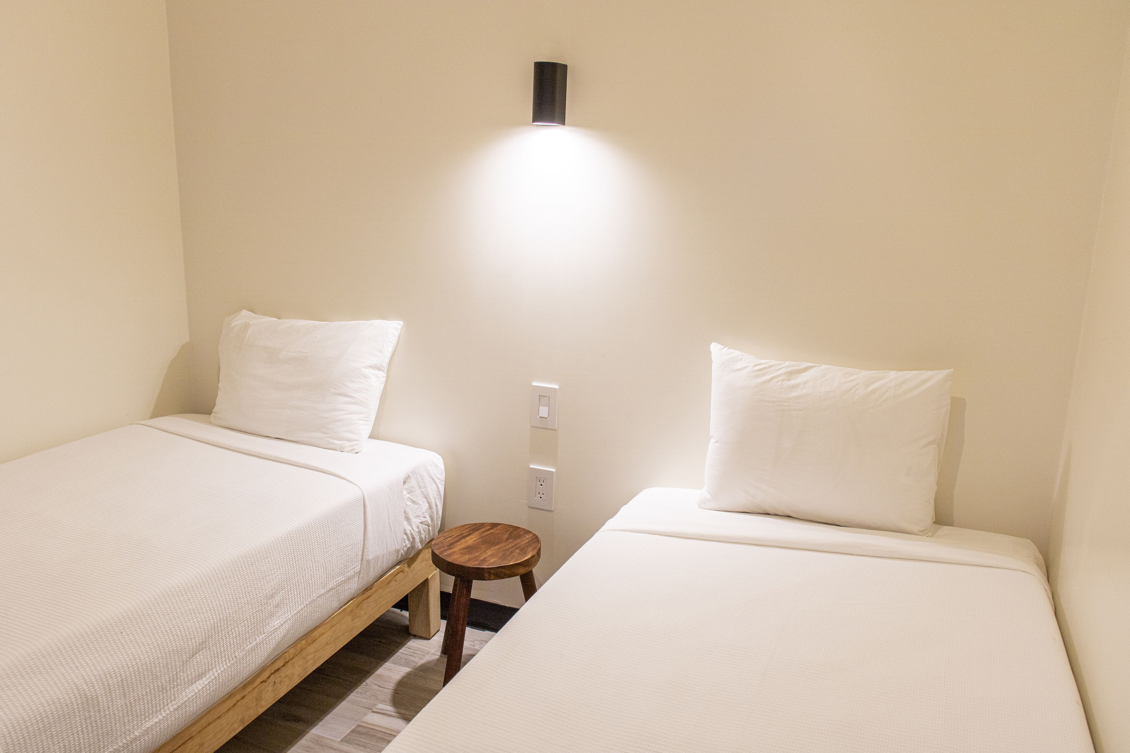A small, clean room with two single beds, a small wooden stool, and a wall-mounted light fixture.