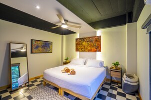 A neatly made bed with white linens, a ceiling fan, and a large abstract painting above it.