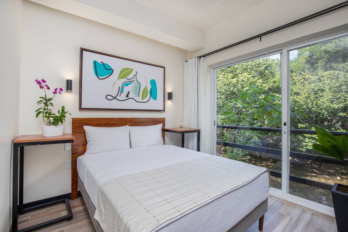 Une chambre dotée d’un grand lit, d’une tête de lit en bois, d’une œuvre d’art accrochée au mur, d’une table de chevet ornée d’une plante et d’une porte-fenêtre coulissante donnant sur un balcon verdoyant.