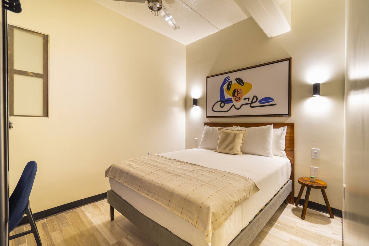 Une chambre d’hôtel avec un lit, une chaise, une petite table et une œuvre d’art accrochée au mur.
