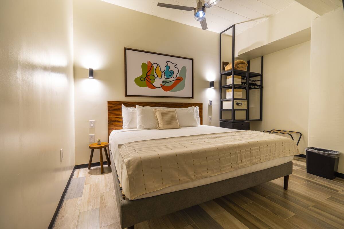 Un lit bien fait dans une chambre d’hôtel, avec une tête de lit en bois, une œuvre d’art accrochée au mur et une petite table de chevet surmontée d’une lampe.