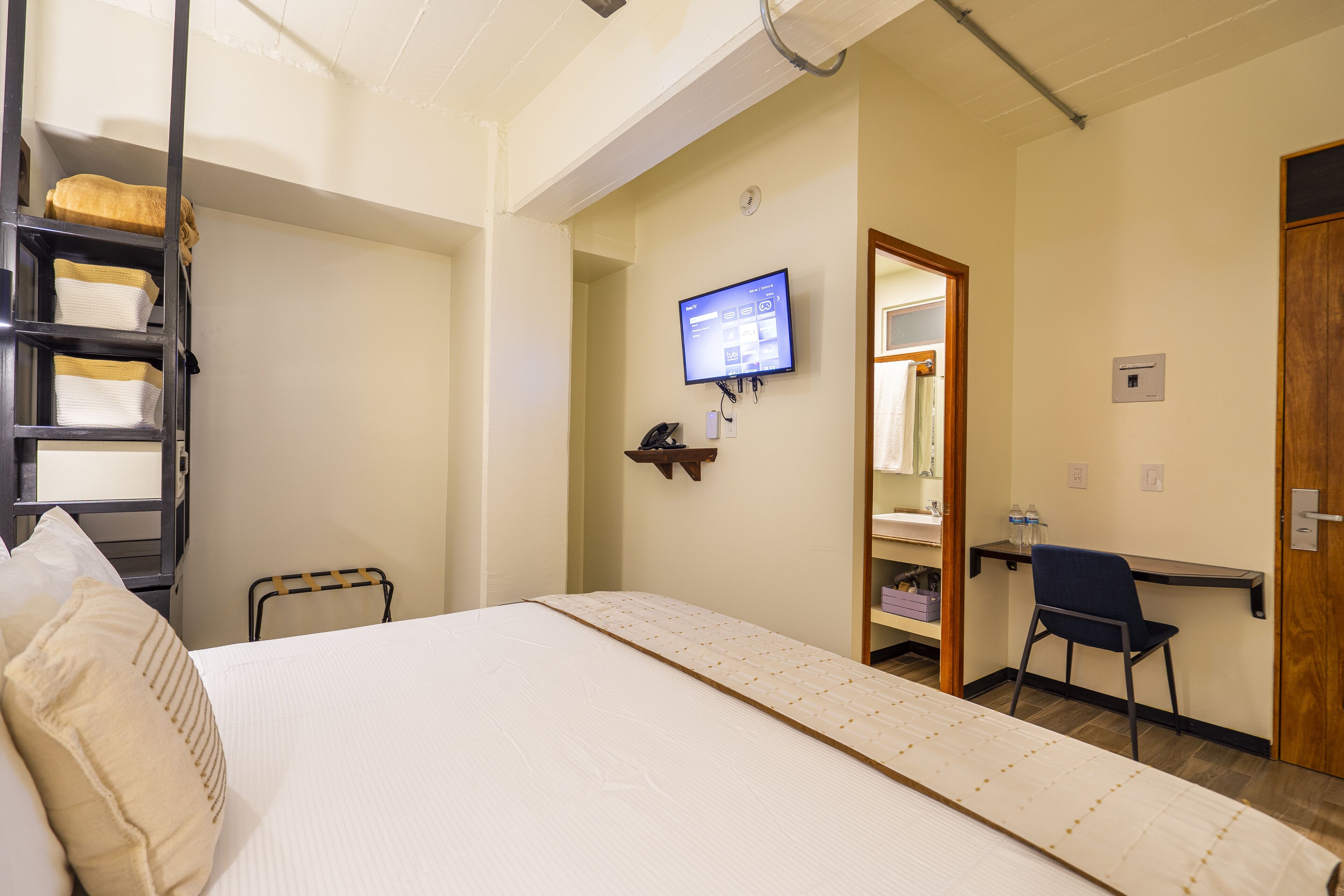 A hotel room with a bed, a desk, a chair, a TV, and a shelf with toiletries.