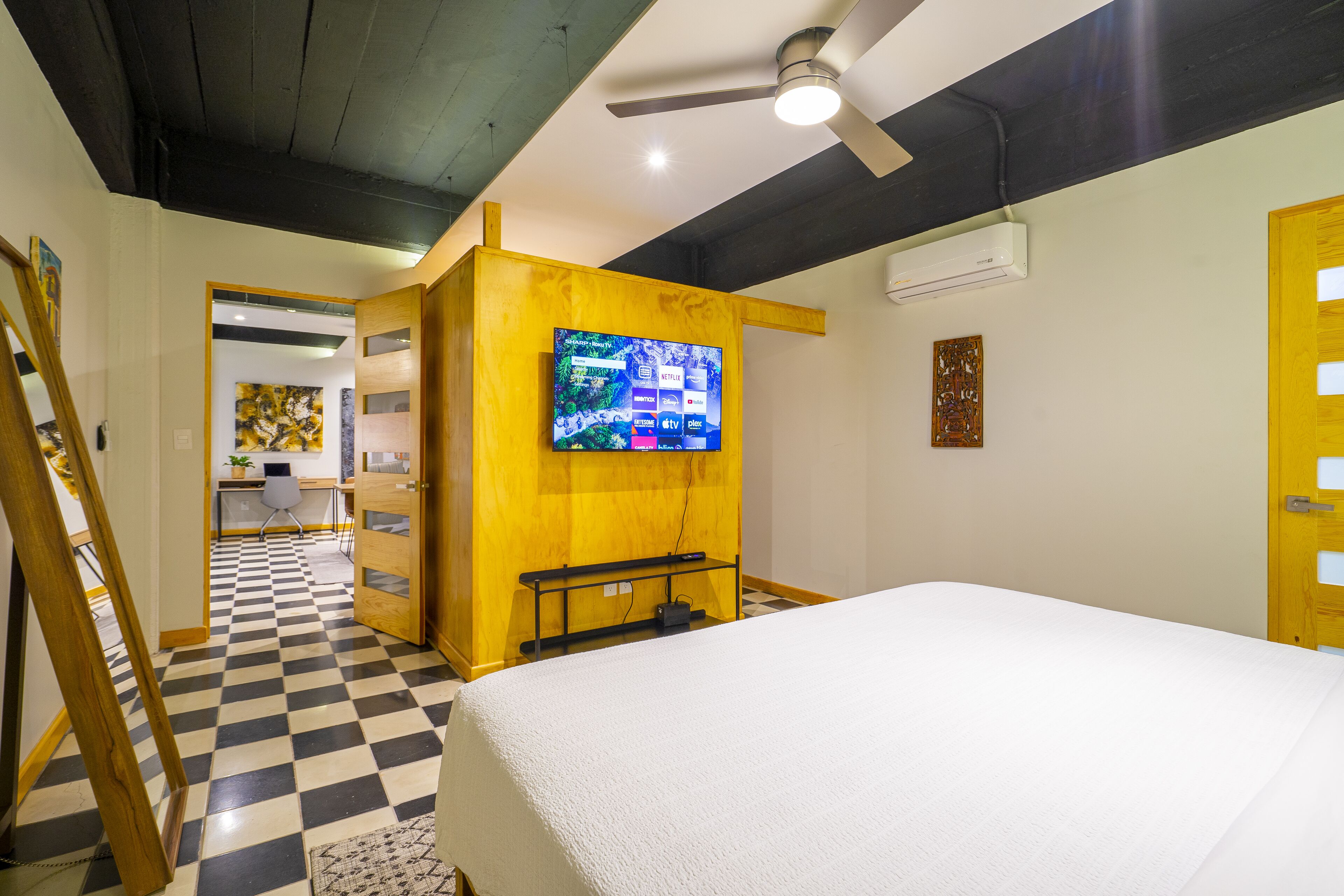 A hotel room with a checkered floor, a bed, a television mounted on a wooden panel, and a ceiling fan.