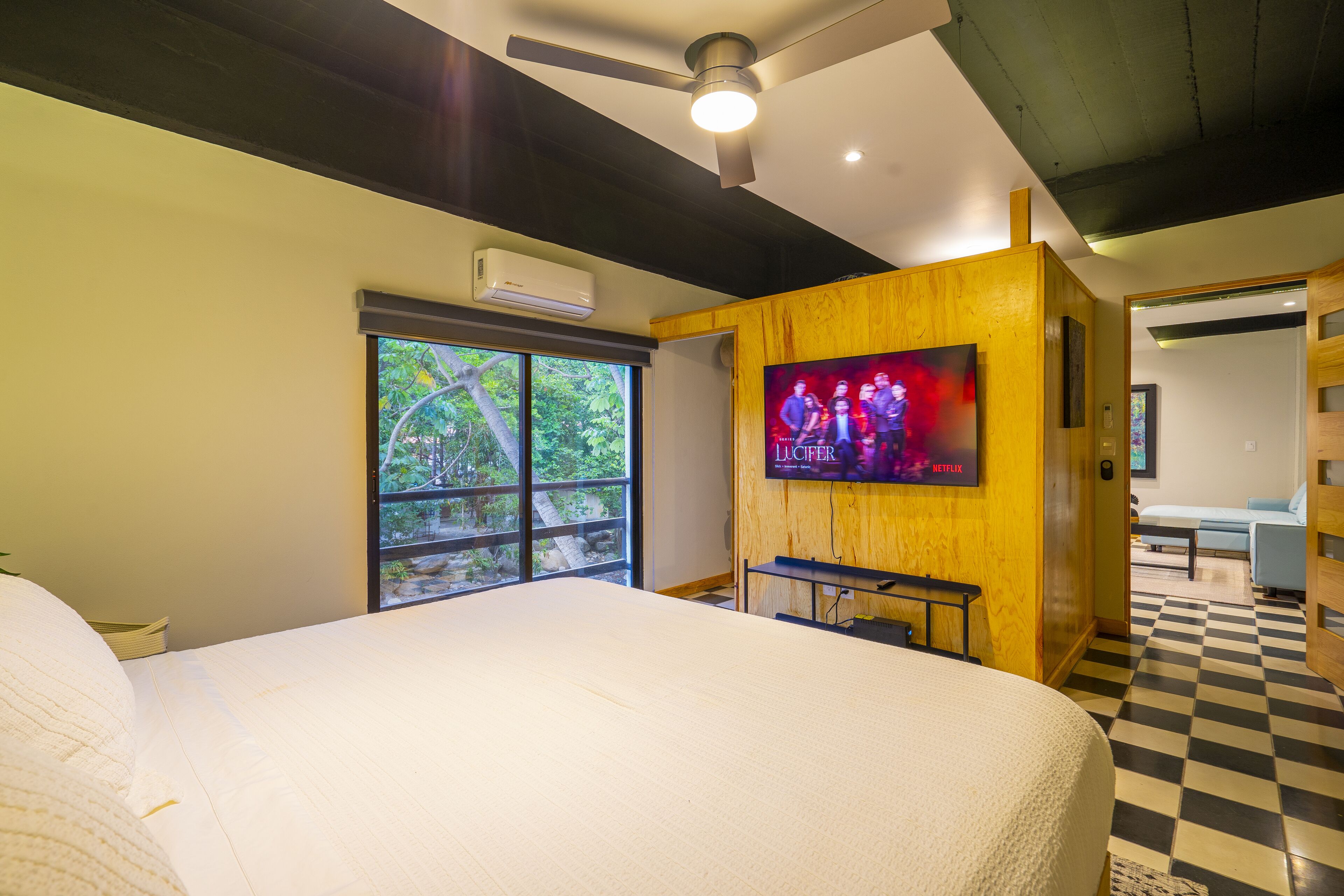 A hotel room with a large bed, a television mounted on the wall, a ceiling fan, and a window with a view of greenery.