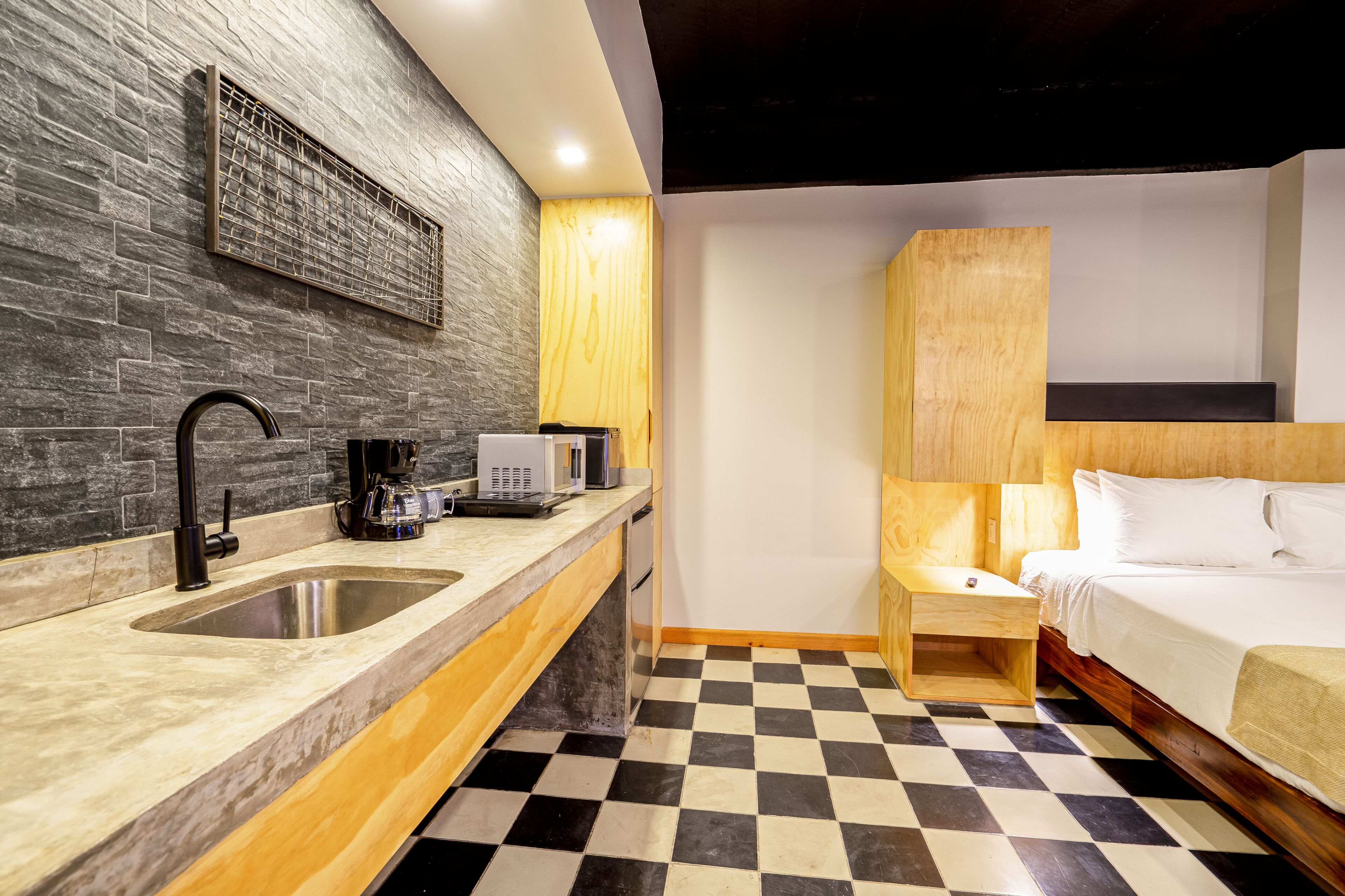 A modern hotel room with a checkered floor, a wooden headboard, a bed, a nightstand, a sink, and a coffee machine.