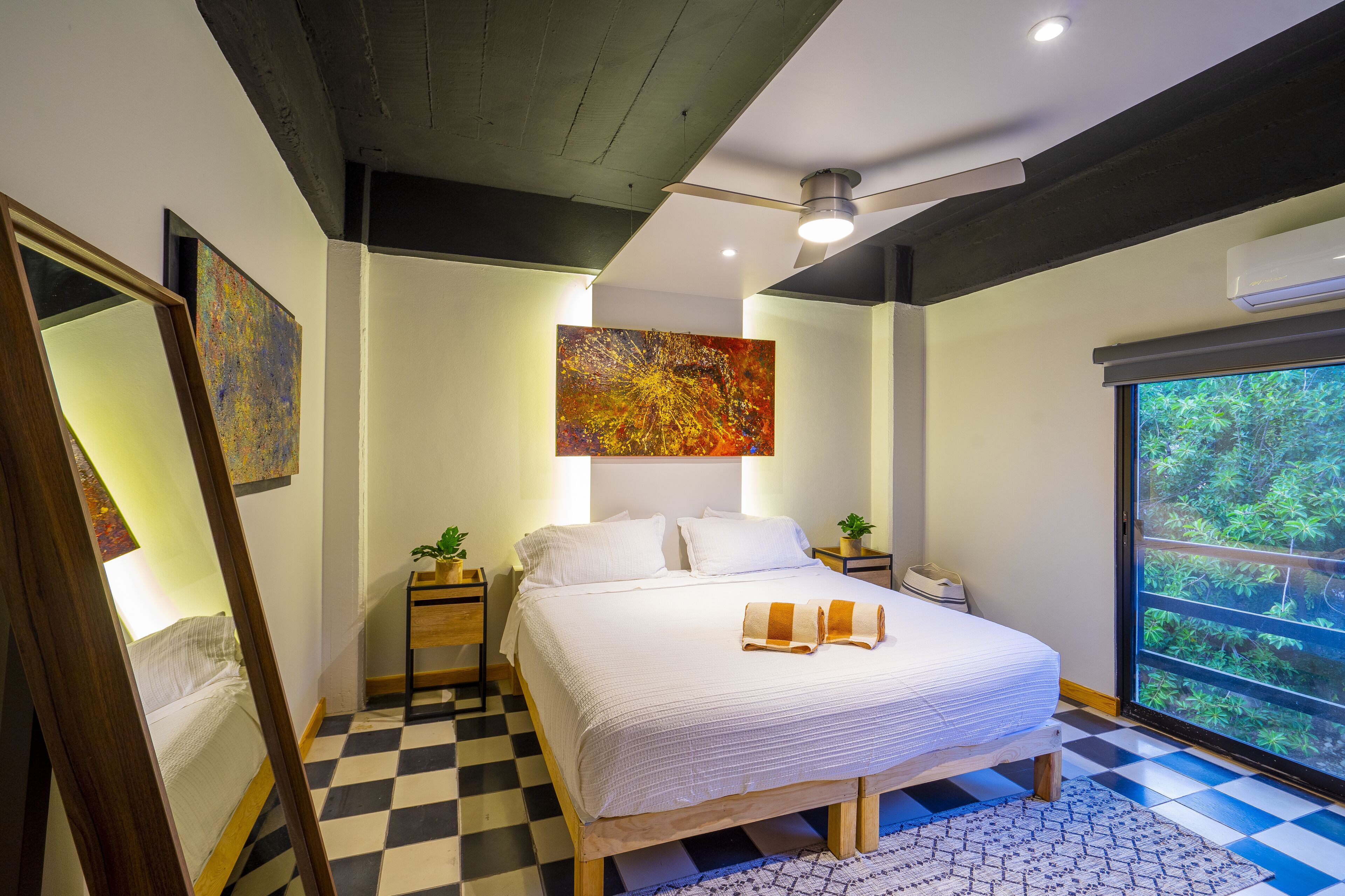 A modern bedroom with a checkered floor, a large bed with white bedding, a ceiling fan, and a large window with a view of greenery.