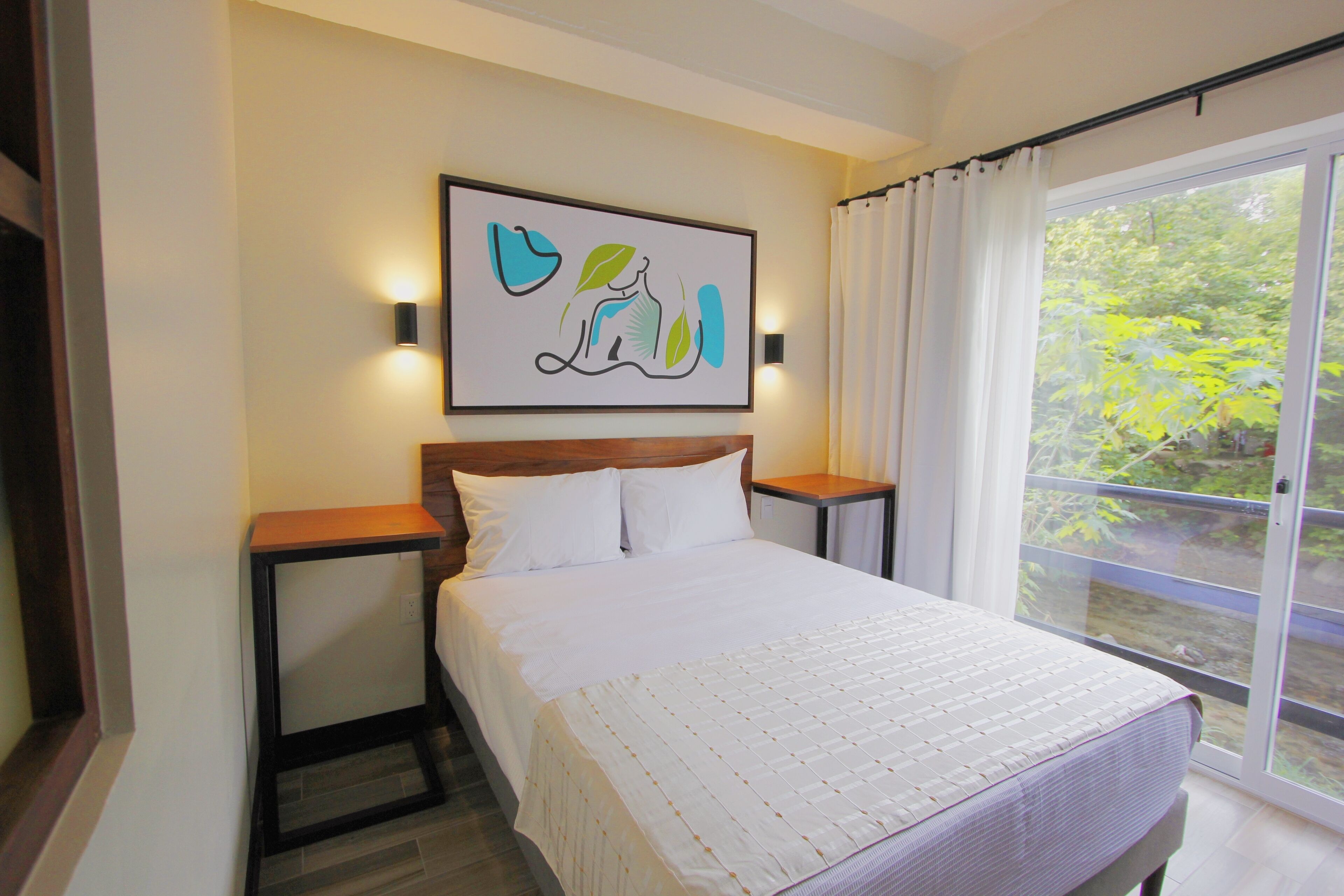 A bedroom with a large bed, a nightstand, a wall-mounted light, and a window with curtains.