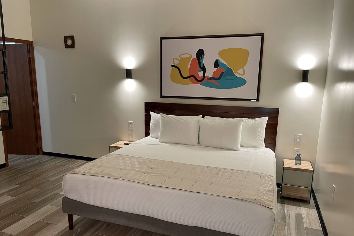 Un lit bien fait, avec du linge de lit blanc, une tête de lit en bois et un plaid beige. Il y a deux tables de chevet avec des lampes, et une œuvre d’art abstraite encadrée est accrochée au-dessus du lit.