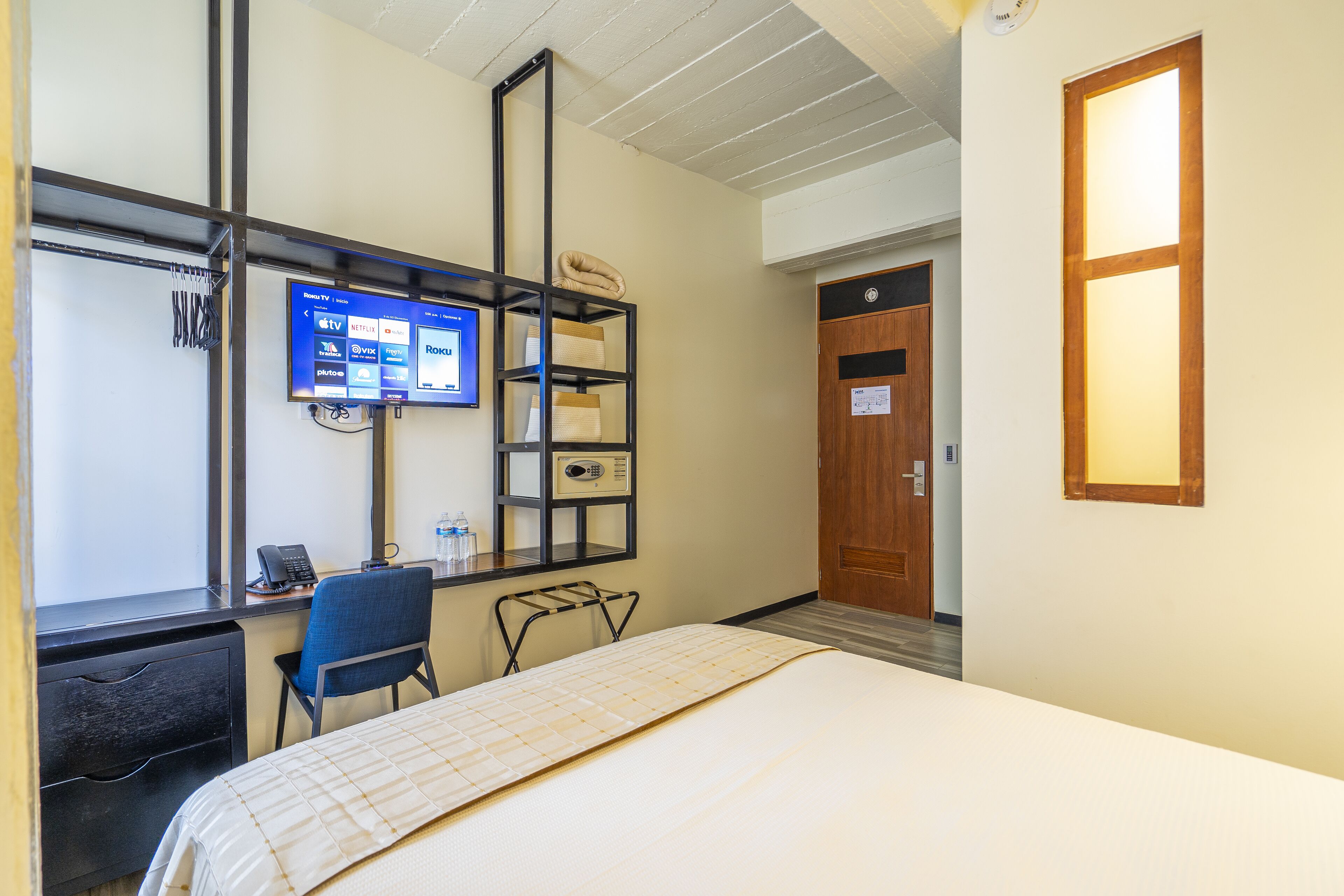 A hotel room with a bed, a desk with a computer, a TV mounted on the wall, and a wooden door.