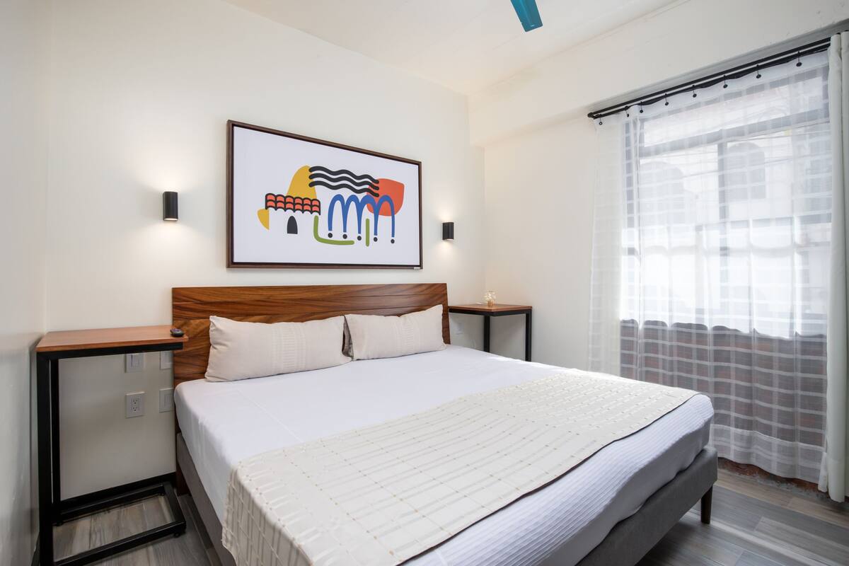 Un lit bien fait, avec une couette blanche et des oreillers, une tête de lit en bois, une table de chevet avec une lampe, et une œuvre d’art abstraite encadrée accrochée au mur.